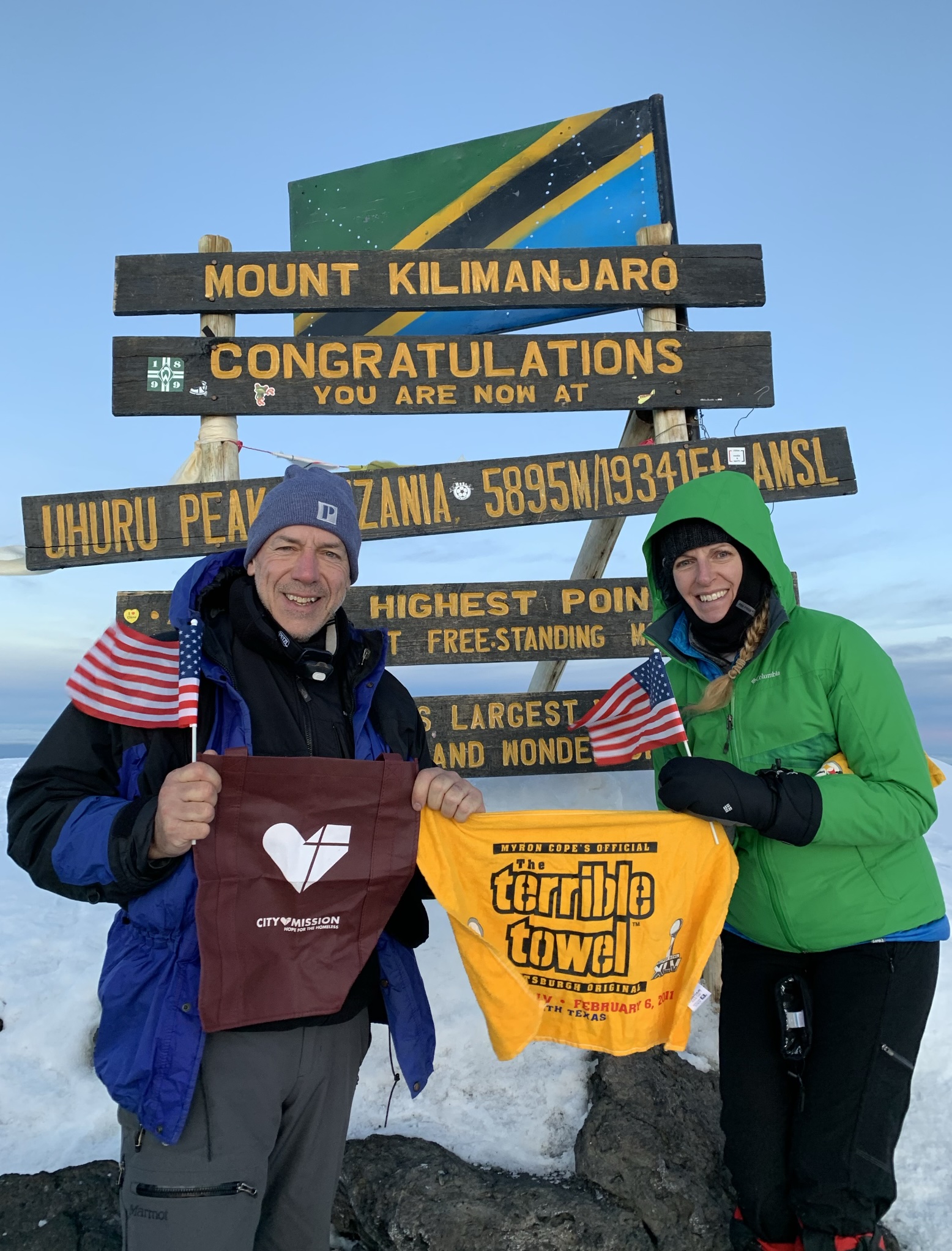 Brenda and Dave at the summit of Mount Kilimanjaro holding an American flag and a Terrible Towel
